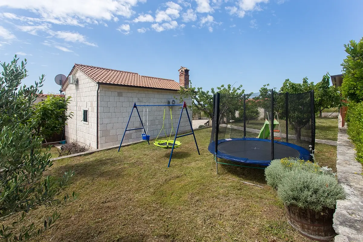 Same private outdoor children's playground in the garden of the house, viewed from a different angle, featuring swings, a slide, and a trampoline.
